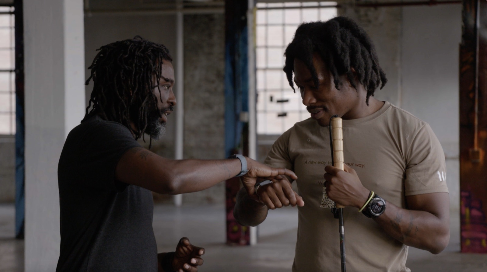 Two men standing together in a bright workshop space. One demonstrates a haptic navigation wristband on his wrist while the other, holding a white cane, reaches out to feel it.